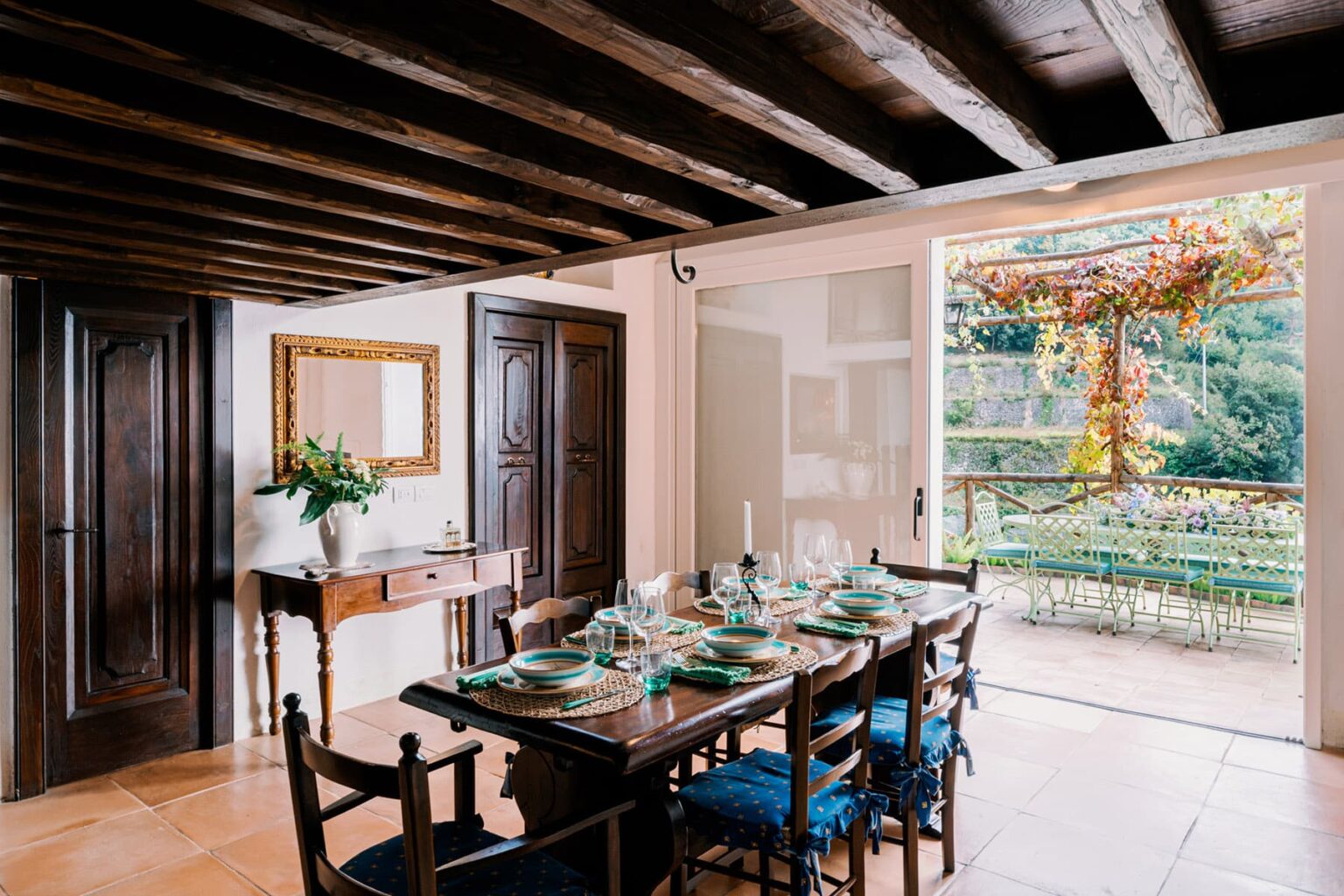 Cozy dining room with a wooden table set for a meal, featuring elegant dishware and a view of a garden through large glass doors, highlighting rustic beams and warm decor. | Only on SopranoVillas