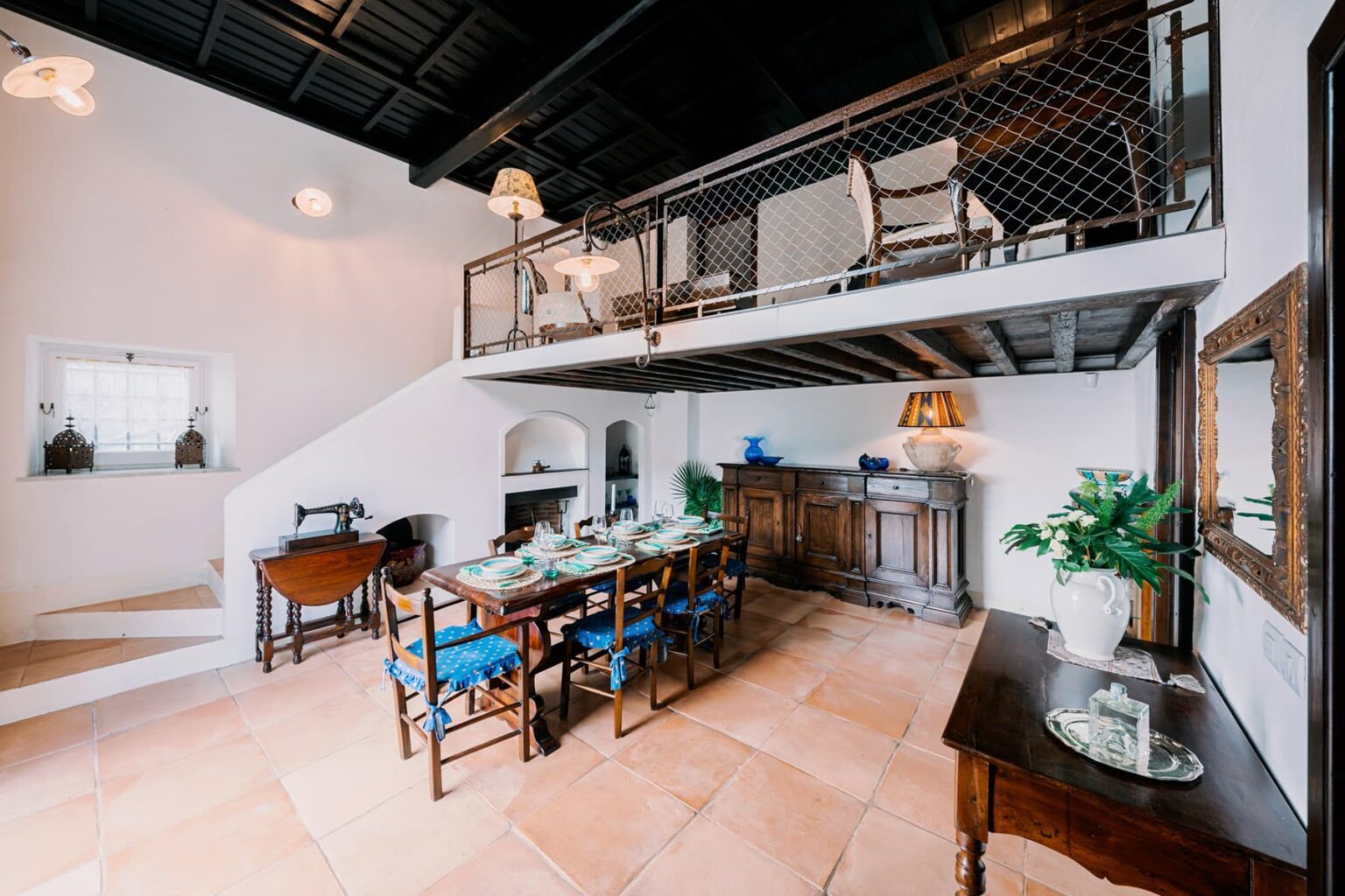 Spacious interior of a modern dining area with a wooden table and blue chairs, featuring a cozy loft above, terracotta flooring, and stylish decor elements. | Only on SopranoVillas