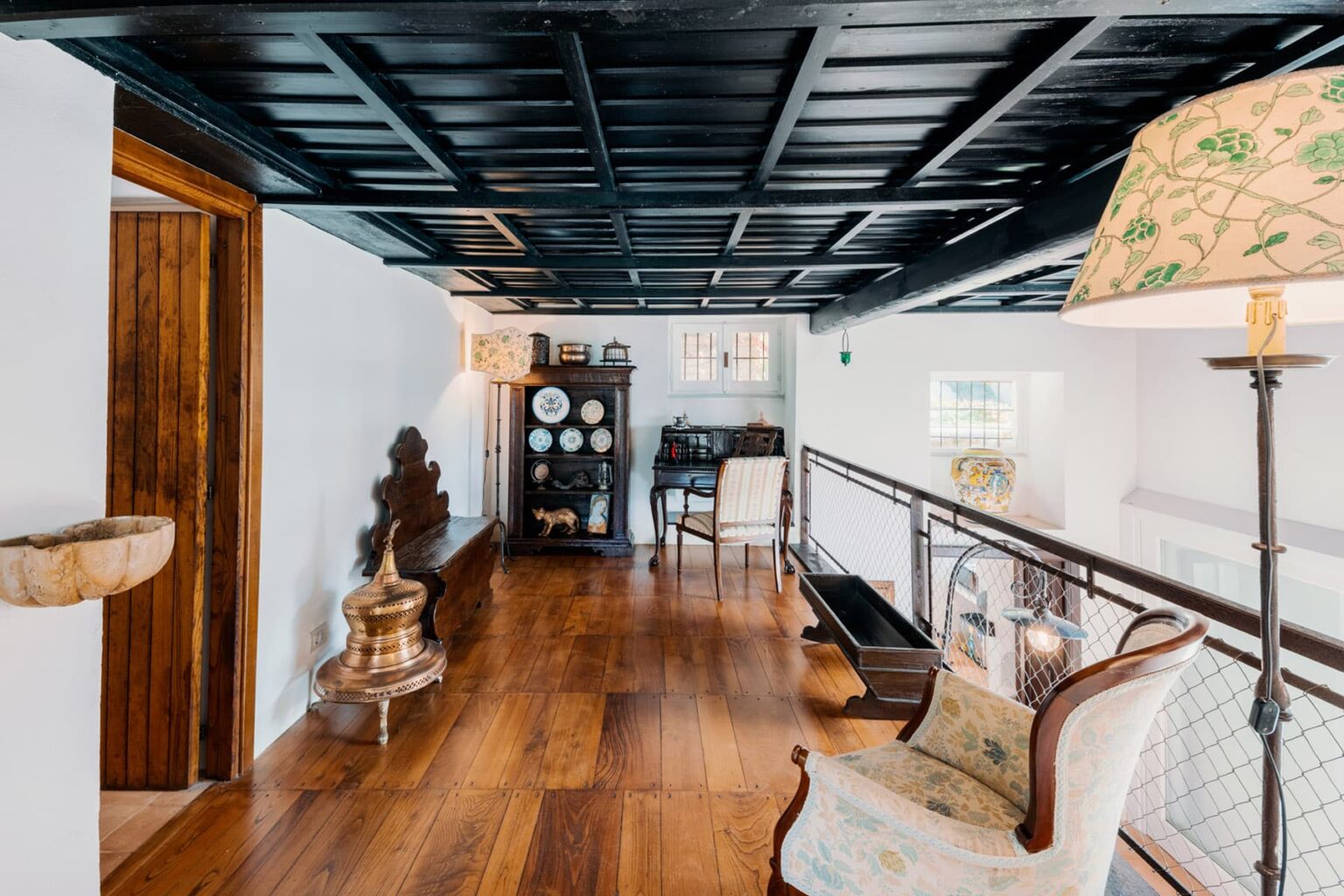 Interior view of a stylish hallway featuring dark coffered ceilings, warm wooden flooring, and vintage decor. The space includes a decorative lamp, an antique cabinet displaying porcelain, and a comfortable armchair, creating an inviting atmosphere. | Only on SopranoVillas