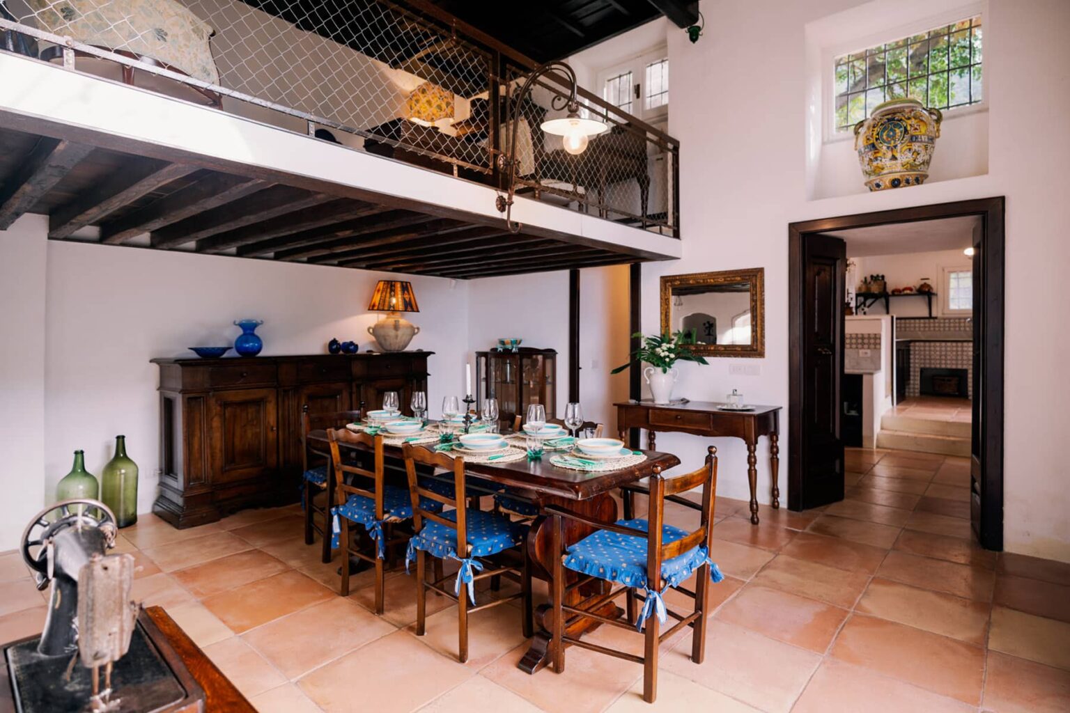 Bright and inviting dining area featuring a wooden table set for a meal, surrounded by blue upholstered chairs. The room showcases rustic decor with a wooden sideboard, large windows allowing natural light, and a glimpse of an open kitchen in the background. | Only on SopranoVillas