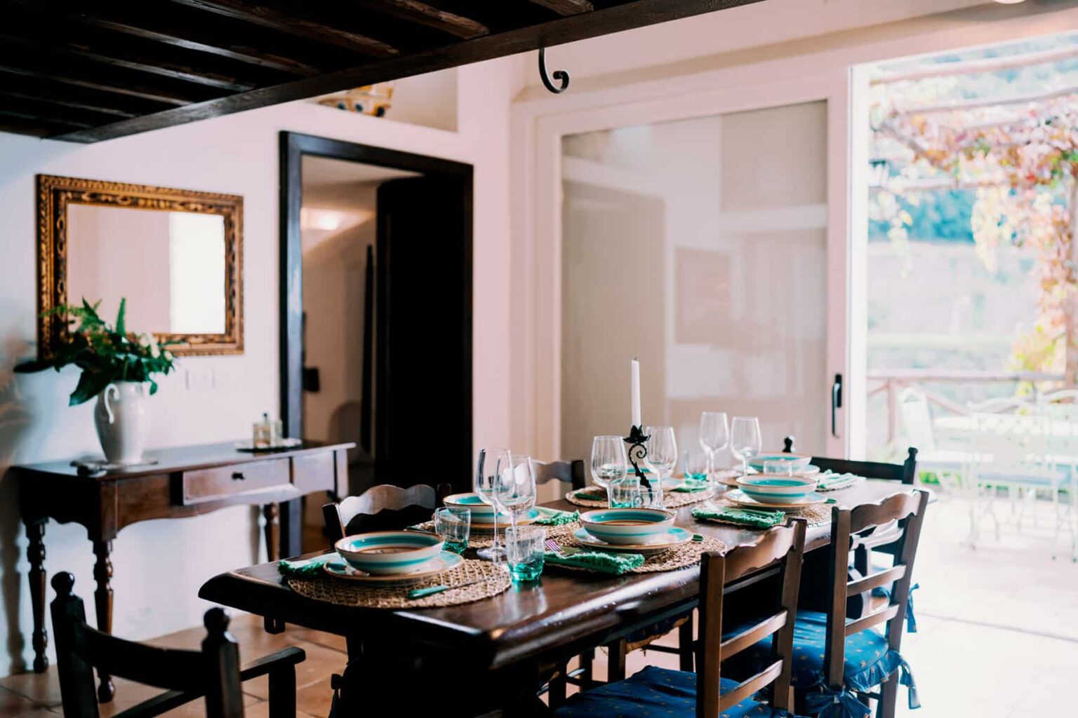 Cozy dining room featuring a wooden table set for a meal with turquoise plates, glassware, and woven placemats. A vintage mirror and a decorative vase with plants adorn the wall, while large glass doors open to a patio with outdoor seating. | Only on SopranoVillas