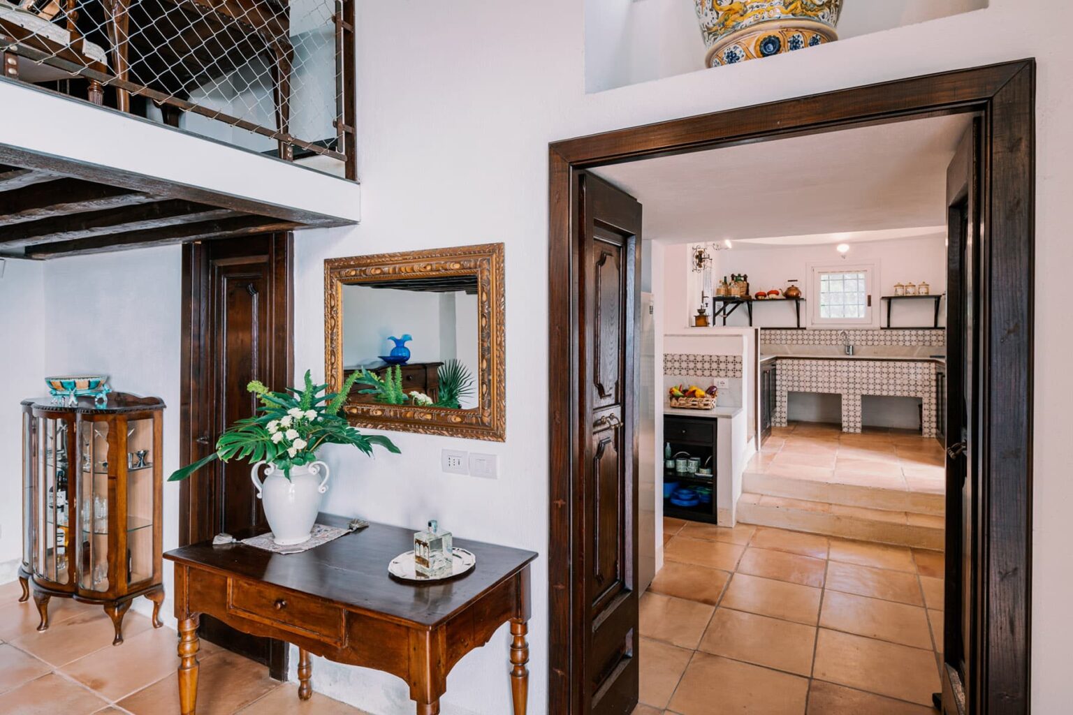 Interior view of a stylish home showcasing an entryway with a wooden table, decorative mirror, and a potted plant, leading to a spacious kitchen area with tiled floors and modern appliances. | Only on SopranoVillas
