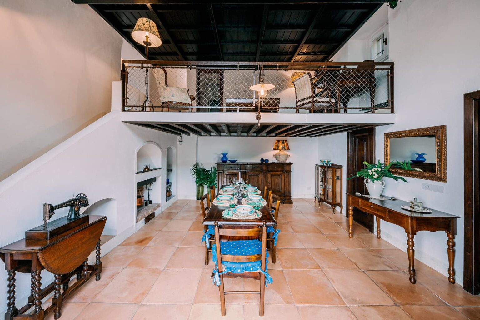 Spacious dining area featuring a long wooden table set for a meal, surrounded by elegant chairs. The room has a rustic charm with terracotta flooring, white walls, and wooden beams. A cozy loft area is visible above, adorned with stylish furnishings and decorative plants. | Only on SopranoVillas