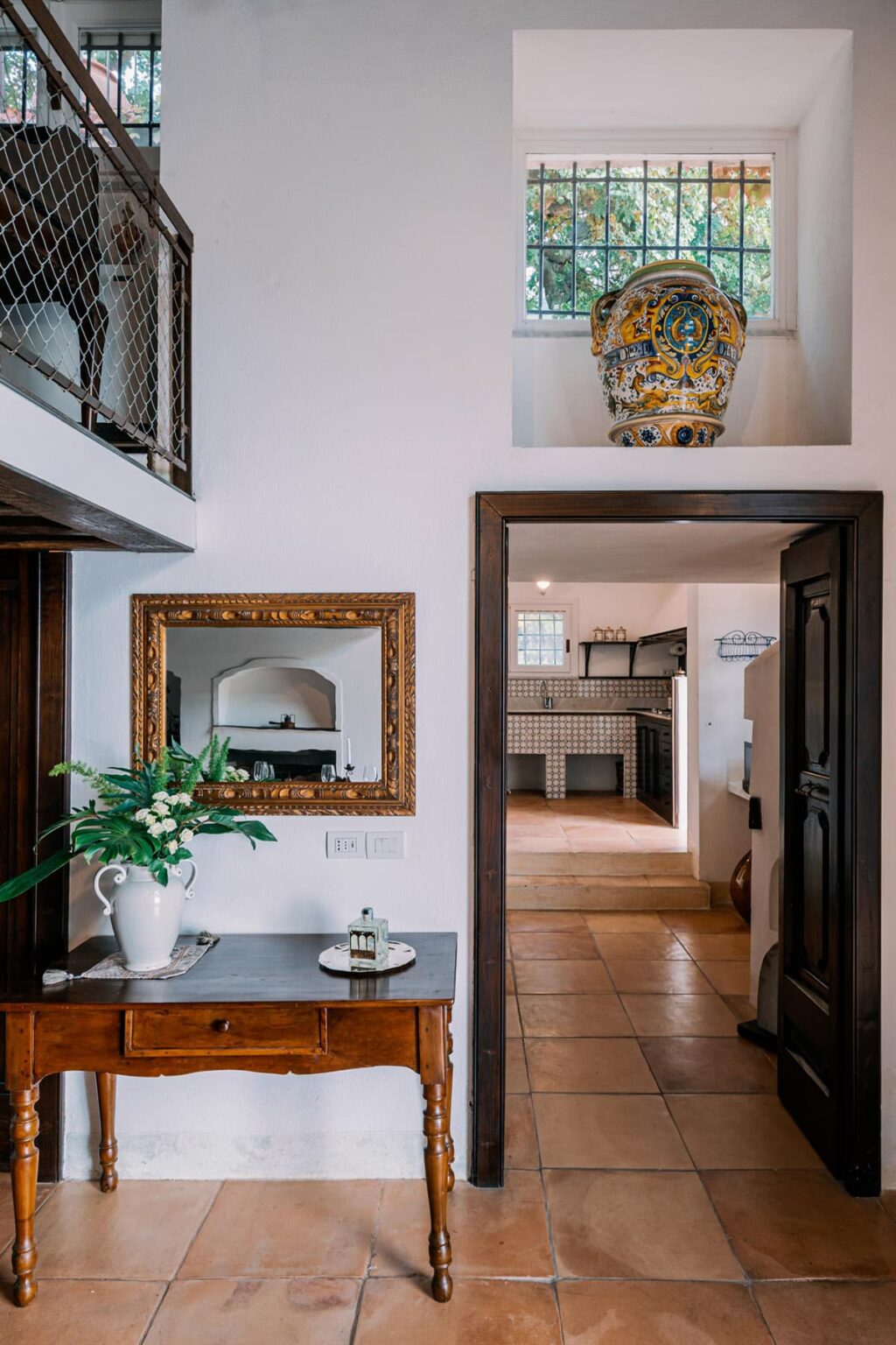 Interior view of a stylish home featuring a decorative mirror in a vintage frame, a vase with flowers on a table, and an open doorway leading to a modern kitchen with tiled backsplash and wooden flooring. | Only on SopranoVillas