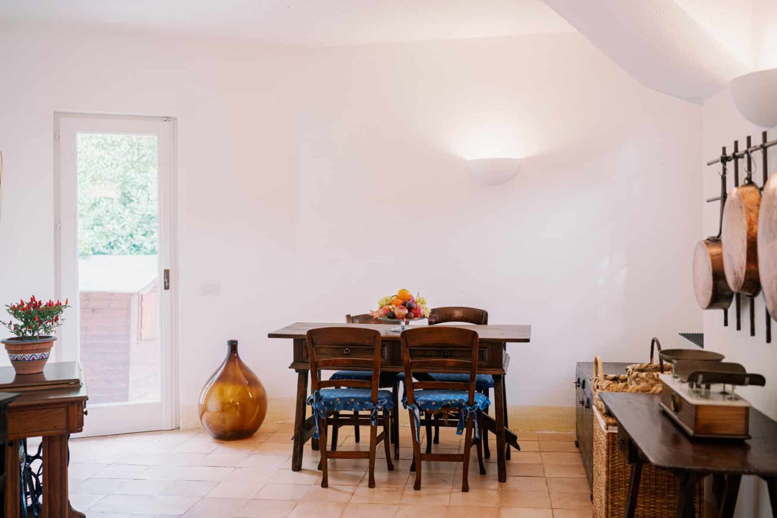 Modern dining area featuring a wooden table with chairs, a decorative vase, and wall-mounted cookware, illuminated by soft lighting and natural light from a door leading outside. | Only on SopranoVillas