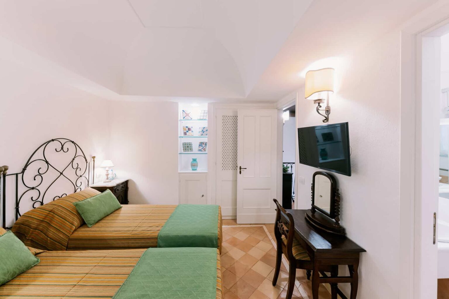 Cozy hotel room featuring two twin beds with green and brown bedding, a wooden desk, and a wall-mounted TV. The room has a bright, modern design with natural light from windows and elegant decor elements. | Only on SopranoVillas