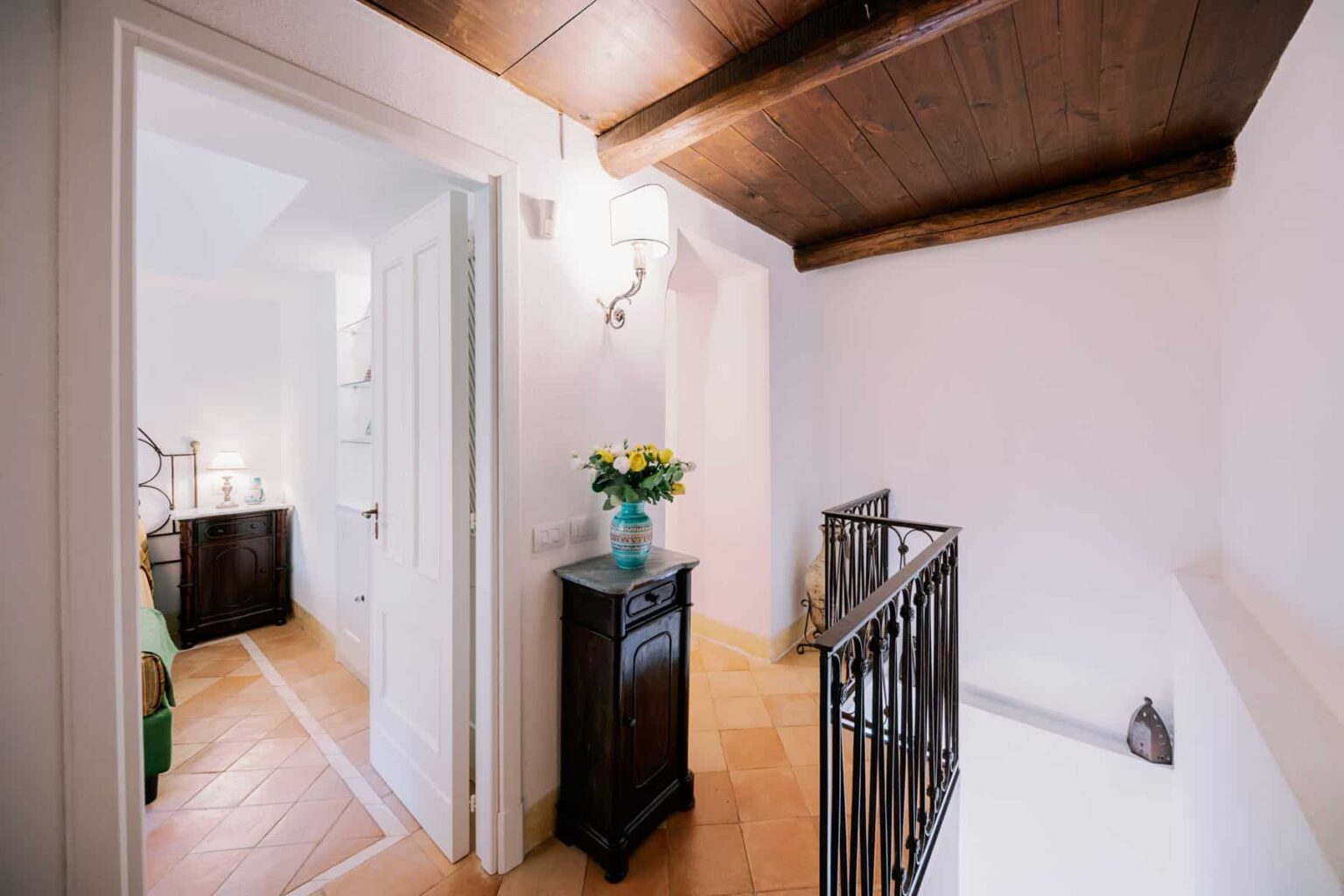 Interior view of a stylish hallway with wooden beams, featuring a small black cabinet and a vase of flowers, leading to a well-lit room. | Only on SopranoVillas
