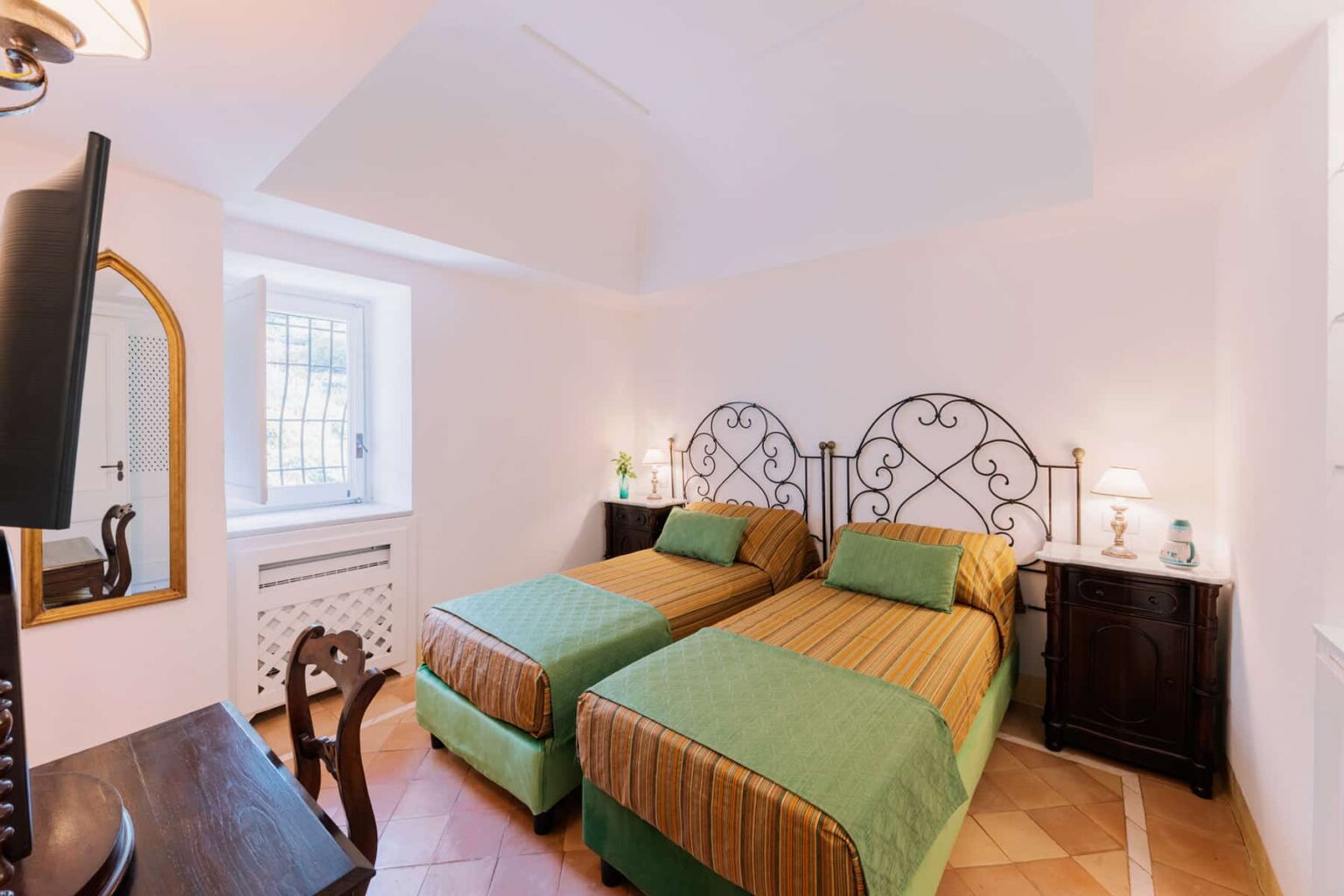 Cozy guest room featuring two twin beds with green and striped bedding, a wooden desk, and a decorative mirror. Natural light streams in through a window, illuminating the room's warm decor and elegant furnishings. | Only on SopranoVillas