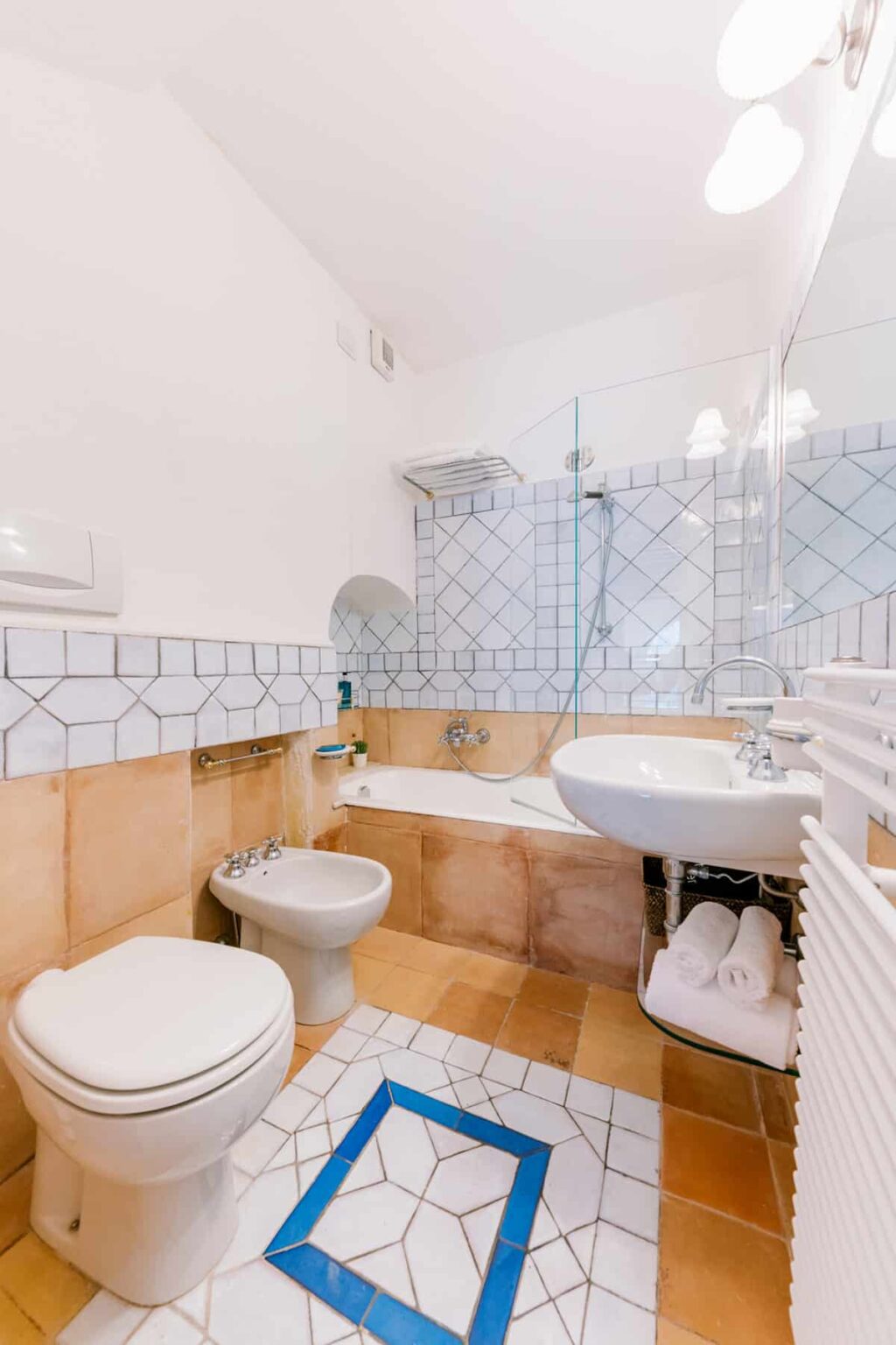 Modern bathroom featuring a white toilet, bidet, and a sleek sink with a mirror. The space includes a glass shower enclosure, a bathtub, and a decorative tiled floor with a blue accent. Soft towels are neatly arranged on a rack, highlighting a clean and inviting atmosphere. | Only on SopranoVillas