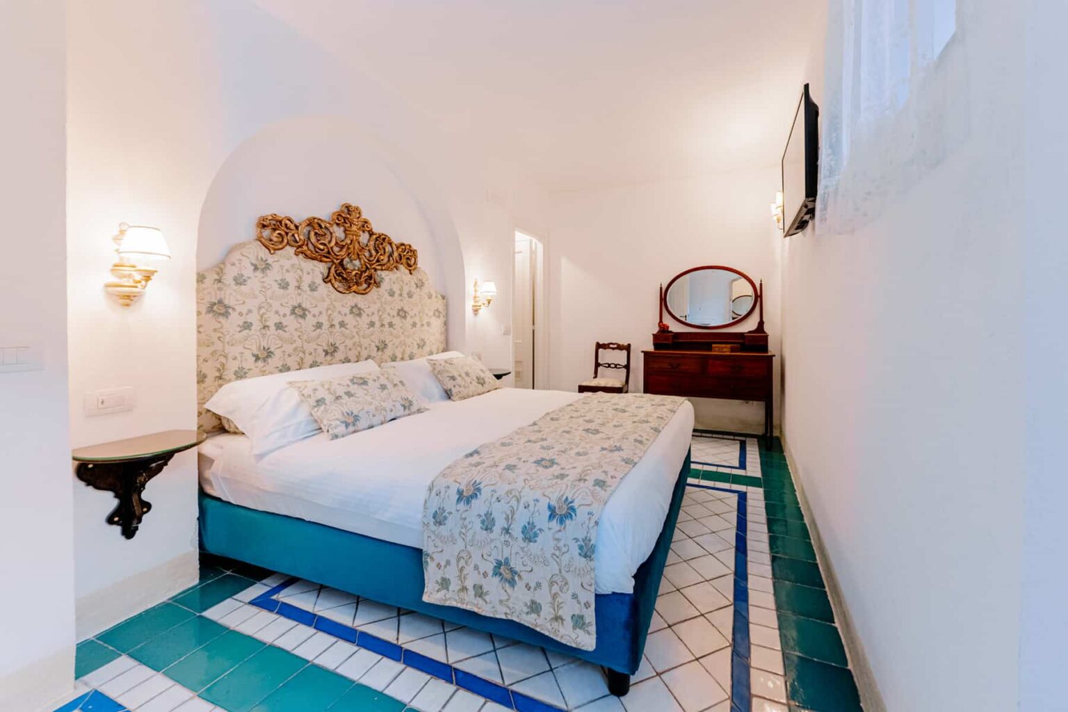 Cozy hotel room featuring two beds with decorative bedding, a vintage mirror, and elegant wall sconces. The room has a unique tiled floor design and a flat-screen TV, providing a stylish and comfortable atmosphere for guests. | Only on SopranoVillas