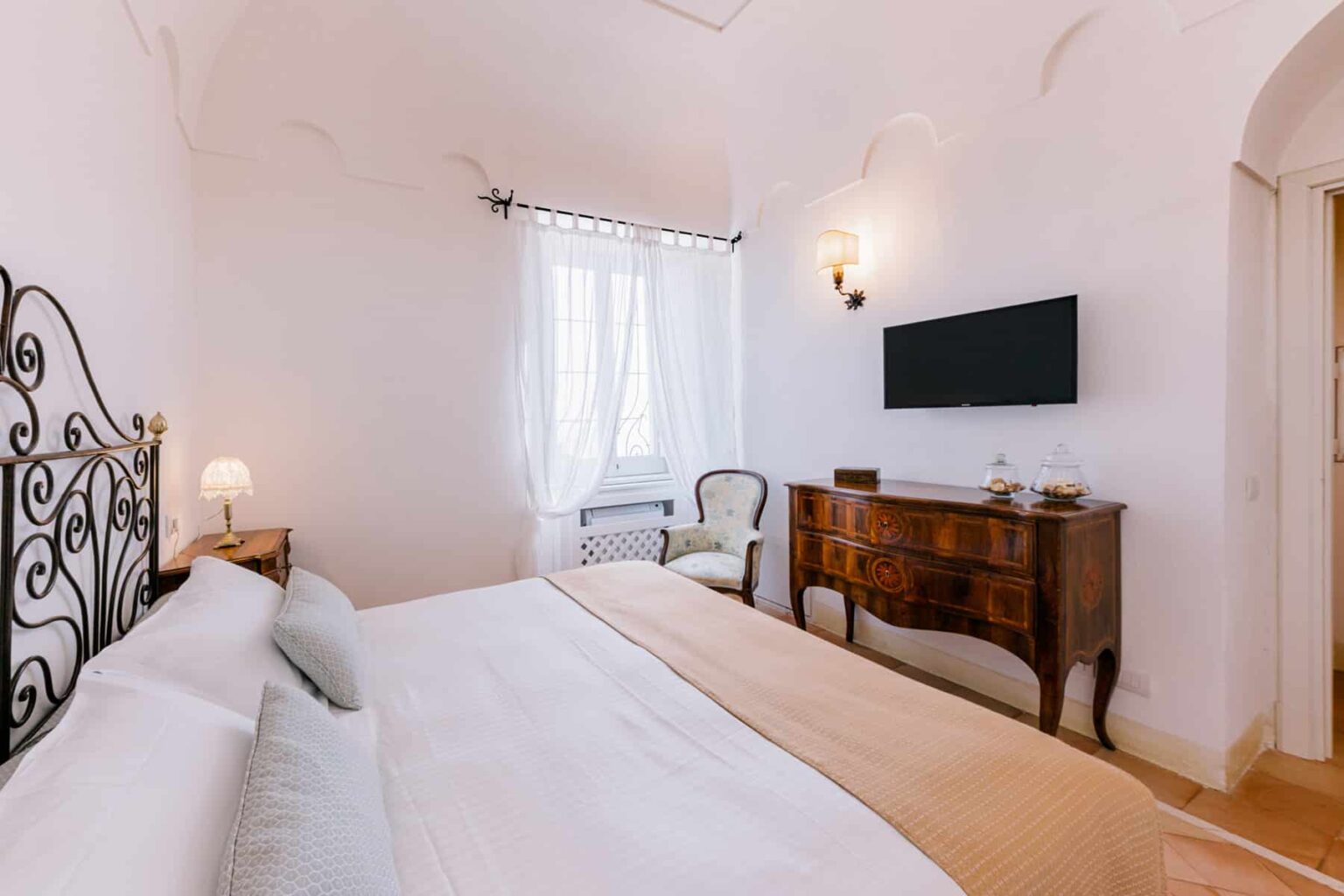 Cozy bedroom featuring a stylish wrought iron bed, elegant wooden dresser, and a modern wall-mounted TV, illuminated by natural light from large windows with sheer curtains. | Only on SopranoVillas