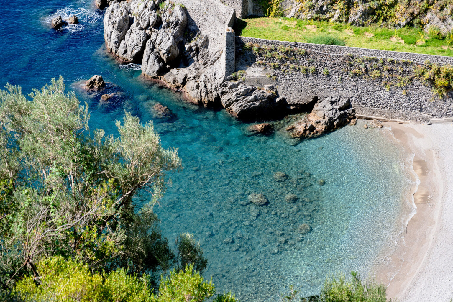 Aerial view of a serene beach with clear turquoise waters, rocky formations, and lush greenery along the shoreline, showcasing a peaceful coastal landscape. | Only on SopranoVillas