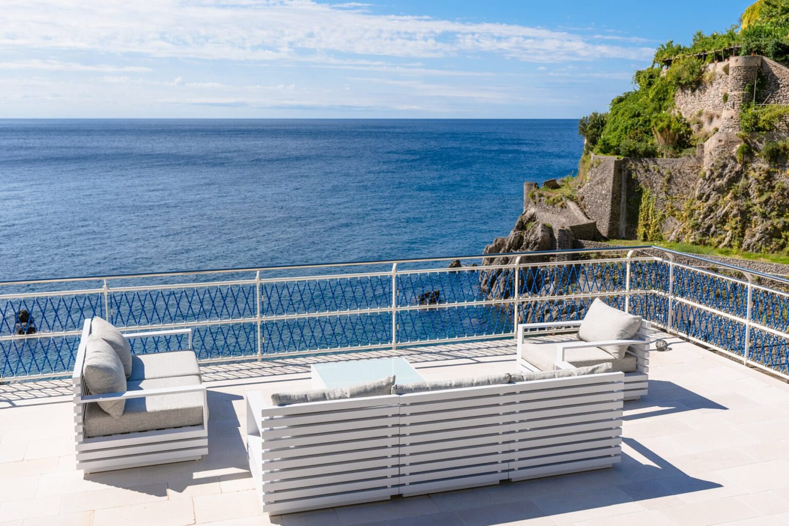 Modern outdoor seating area overlooking a serene ocean view, featuring white furniture and a coastal landscape with cliffs in the background. | Only on SopranoVillas