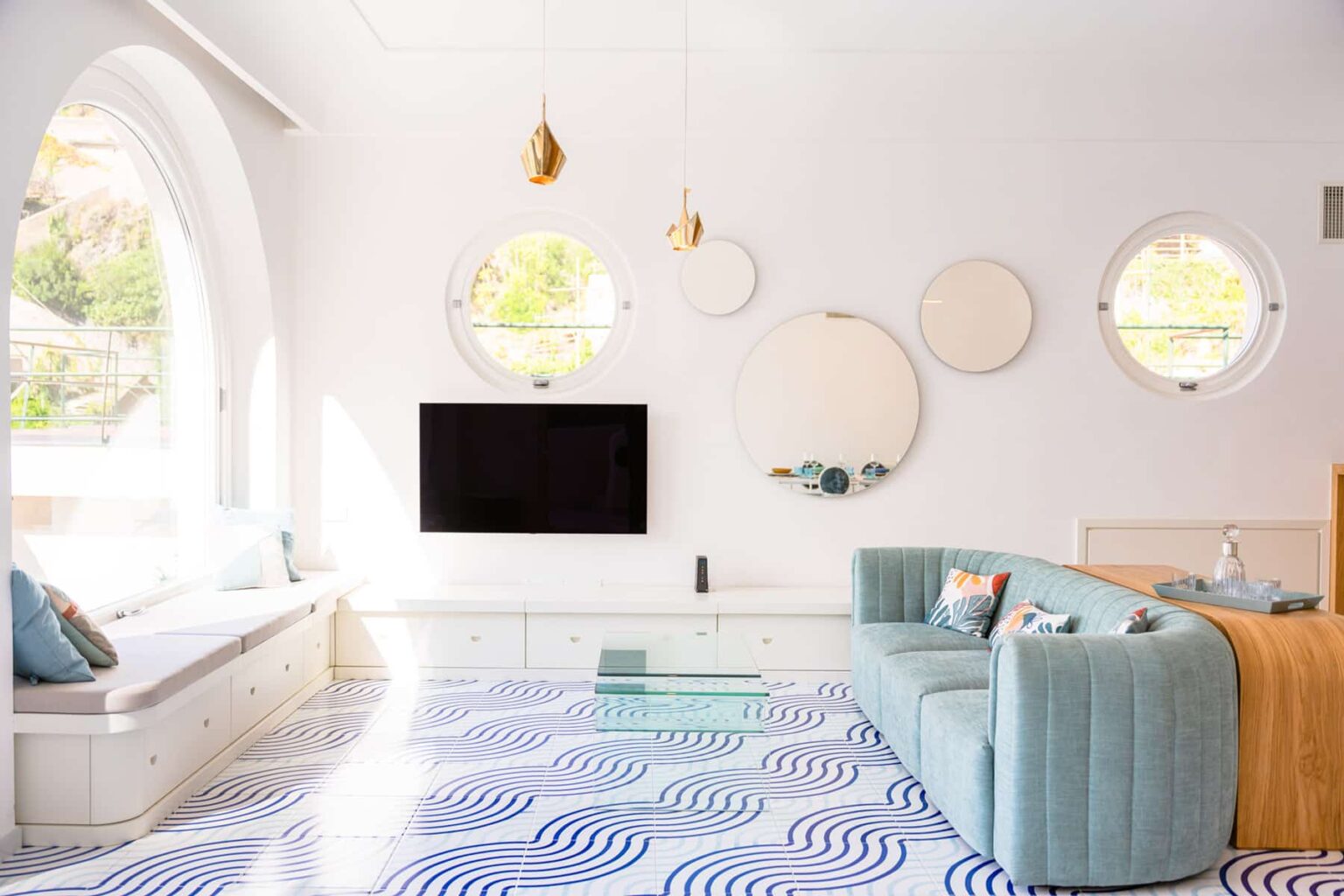 Modern living room featuring a light blue sofa, circular mirrors, and a large flat-screen TV. The space has white walls and a patterned blue and white floor, with natural light streaming in through round windows. Stylish decor elements create a fresh and inviting atmosphere. | Only on SopranoVillas