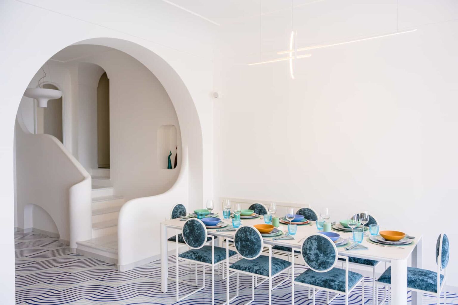 Modern dining room with a white color scheme, featuring a long table set with colorful dishes and teal upholstered chairs. An arched doorway leads to a staircase, enhancing the open and airy feel of the space. | Only on SopranoVillas