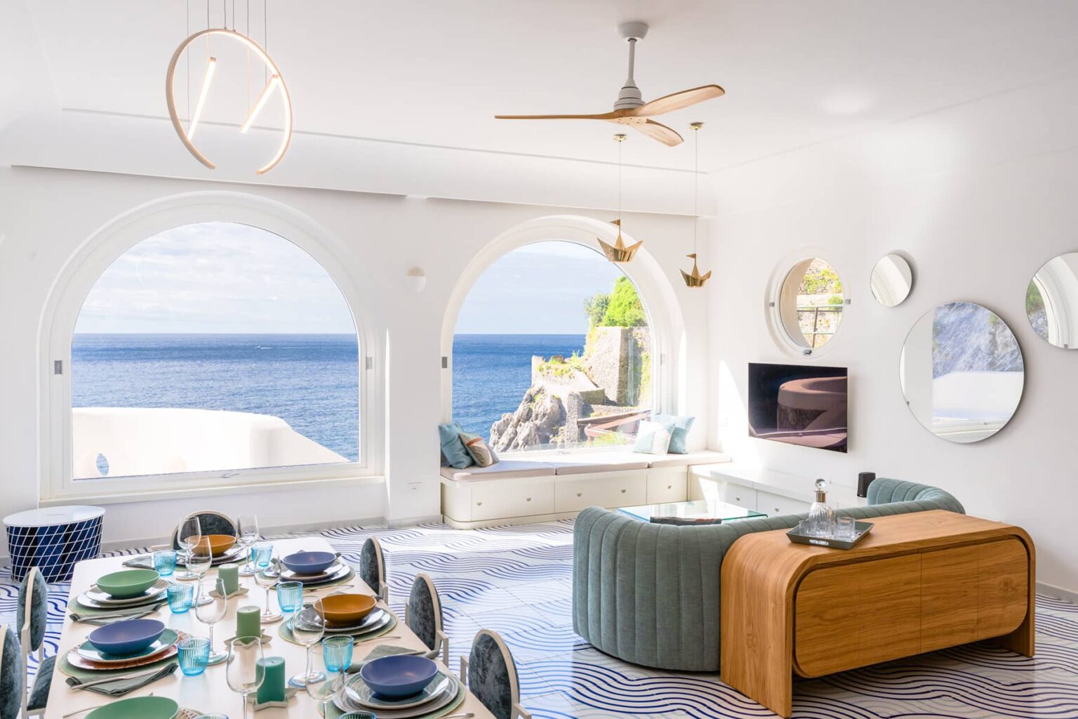 Modern coastal living room with ocean views, featuring an elegant dining table set, a stylish curved sofa, decorative mirrors, and a ceiling fan. Bright natural light illuminates the space through large arched windows. | Only on SopranoVillas