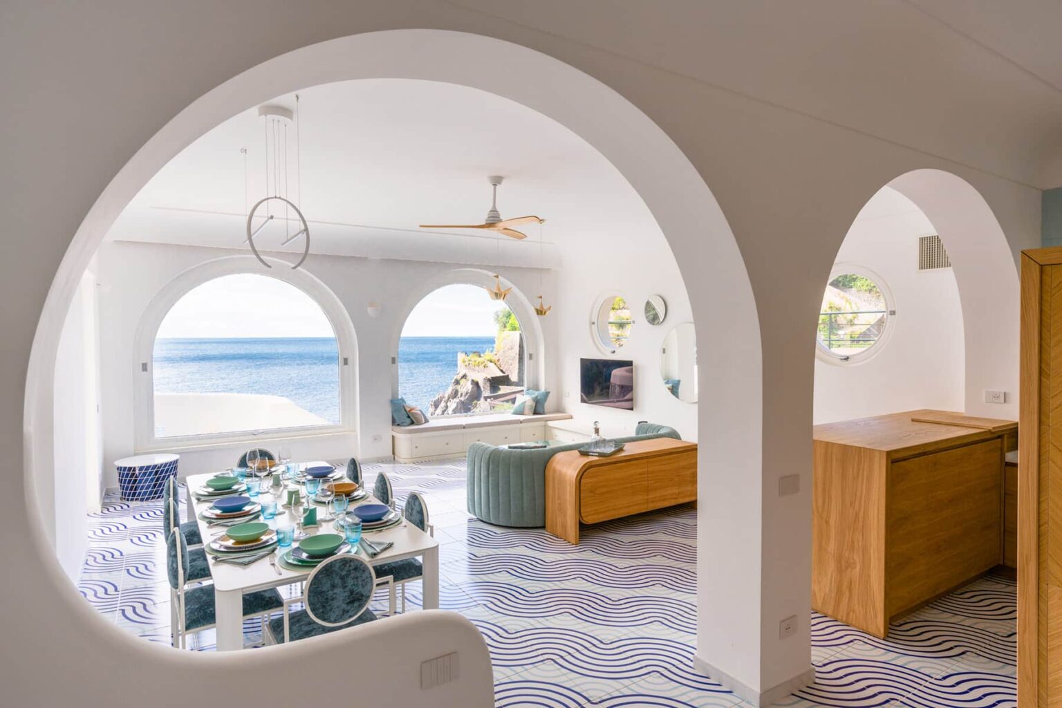 Bright and airy coastal living space with arched doorways, featuring a dining table set with colorful tableware, a modern sofa, and a stunning ocean view through large windows. | Only on SopranoVillas