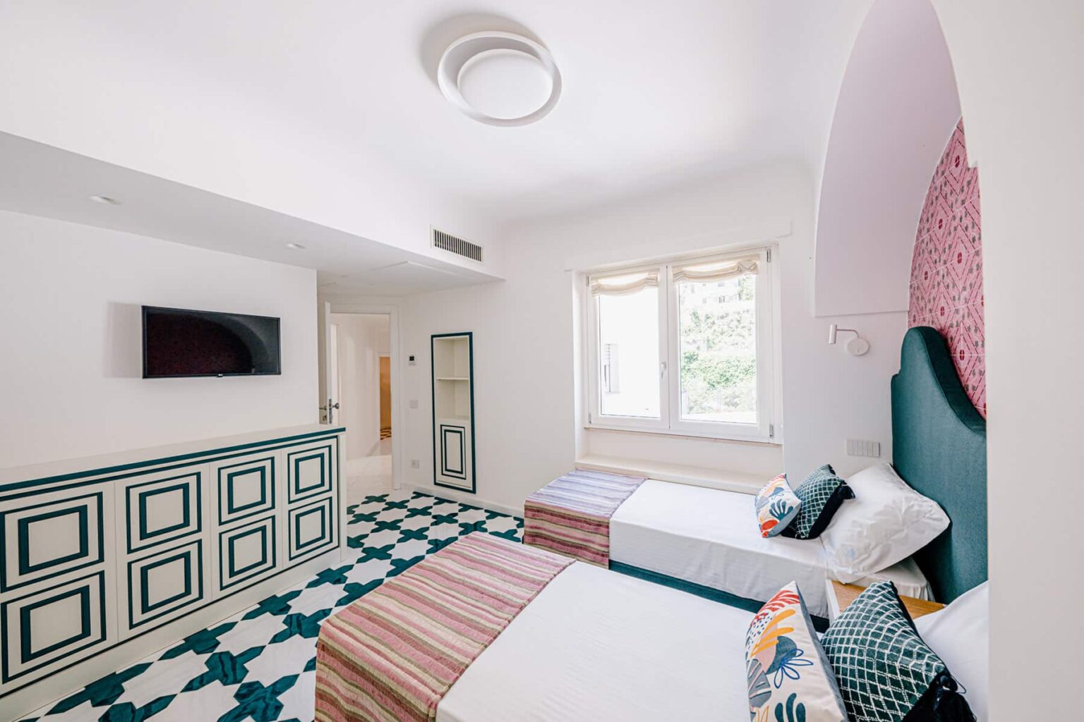 Bright and modern hotel room featuring two twin beds with colorful decorative pillows, a flat-screen TV mounted on the wall, and a stylish patterned floor. Natural light streams in through large windows, enhancing the inviting atmosphere of the space. | Only on SopranoVillas
