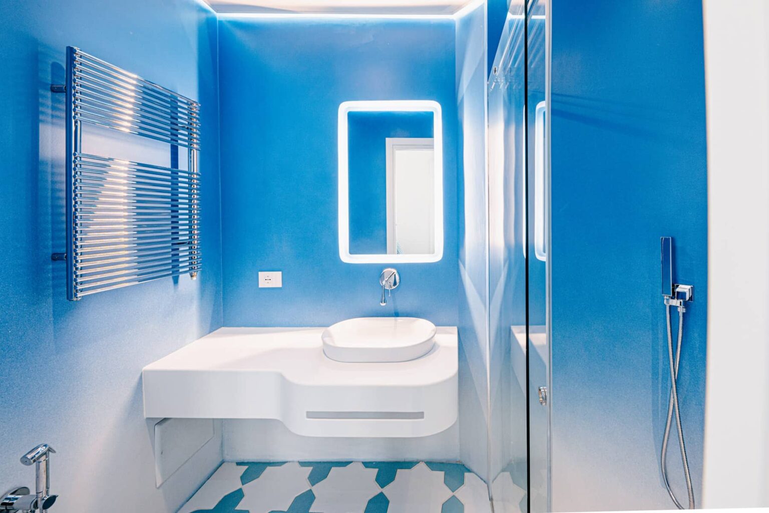 Modern bathroom design featuring bright blue walls, a sleek white countertop with an integrated sink, a backlit mirror, and a glass shower enclosure. The floor is adorned with patterned tiles, enhancing the contemporary aesthetic. | Only on SopranoVillas