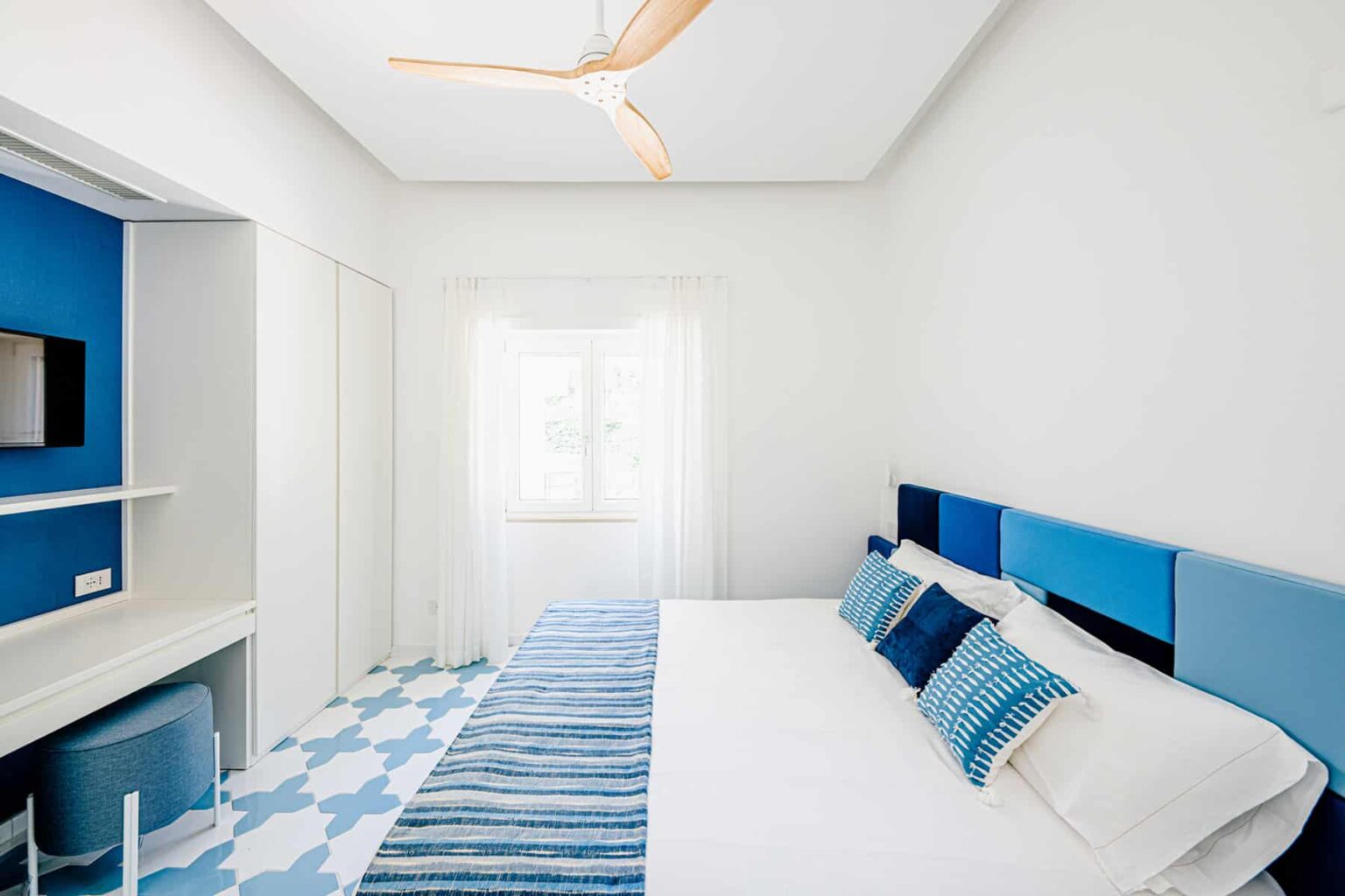 Modern bedroom featuring a white and blue color scheme, a large bed with decorative pillows, a ceiling fan, and a window allowing natural light. The room includes a minimalist design with a television and stylish patterned flooring. | Only on SopranoVillas