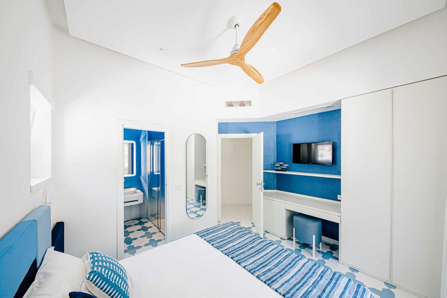 Modern bedroom with blue accents, featuring a white bed with striped blue bedding, a ceiling fan, a flat-screen TV, and a stylish mirror. The room has white walls and a tiled floor, creating a bright and airy atmosphere. | Only on SopranoVillas