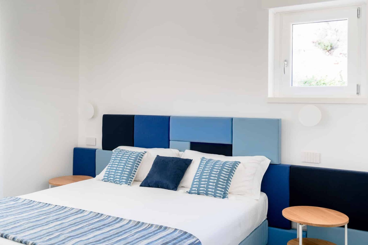 Modern bedroom featuring a stylish blue and white color scheme, a comfortable bed with decorative pillows, and sleek wooden side tables. Natural light fills the space through a window, creating a bright and inviting atmosphere. | Only on SopranoVillas