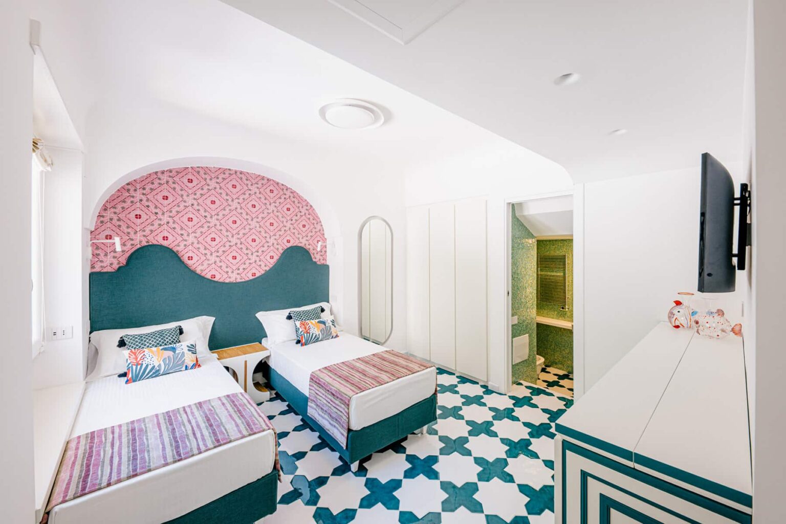 Bright and modern hotel room featuring two twin beds with colorful bedding, a decorative pink wall, patterned tile flooring, and an en-suite bathroom. Ideal for guests seeking a stylish and comfortable stay. | Only on SopranoVillas