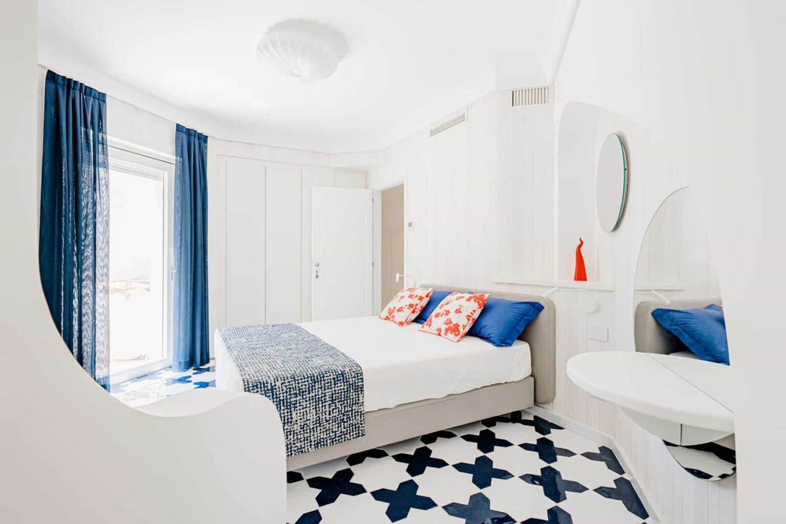 Bright and modern bedroom featuring a double bed with colorful pillows, white walls, and blue curtains. The room includes a stylish round mirror and a unique patterned floor, creating a spacious and inviting atmosphere. | Only on SopranoVillas