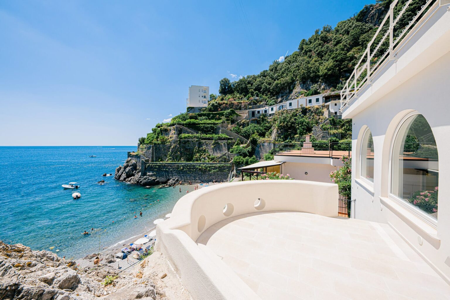 Scenic view of a coastal property overlooking a clear blue sea, with boats anchored near the shore and a lush green hillside in the background. | Only on SopranoVillas