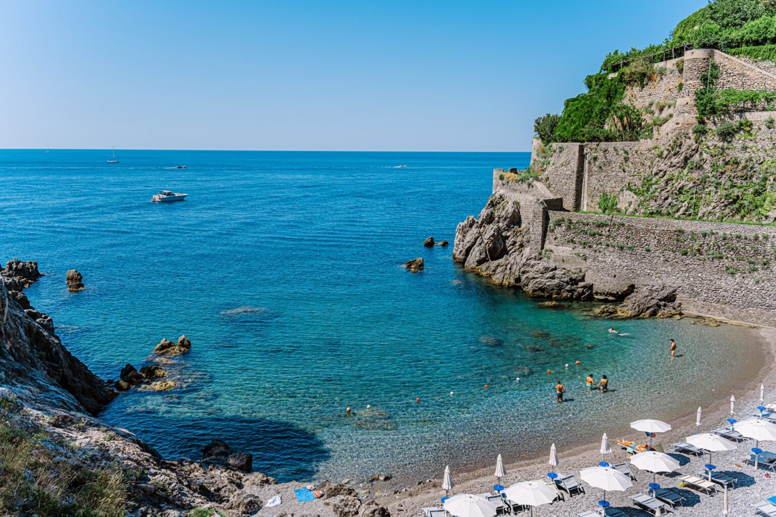 Scenic coastal view featuring a sandy beach with sun loungers, clear turquoise waters, and rocky cliffs under a bright blue sky. Ideal destination for relaxation and beach activities. | Only on SopranoVillas