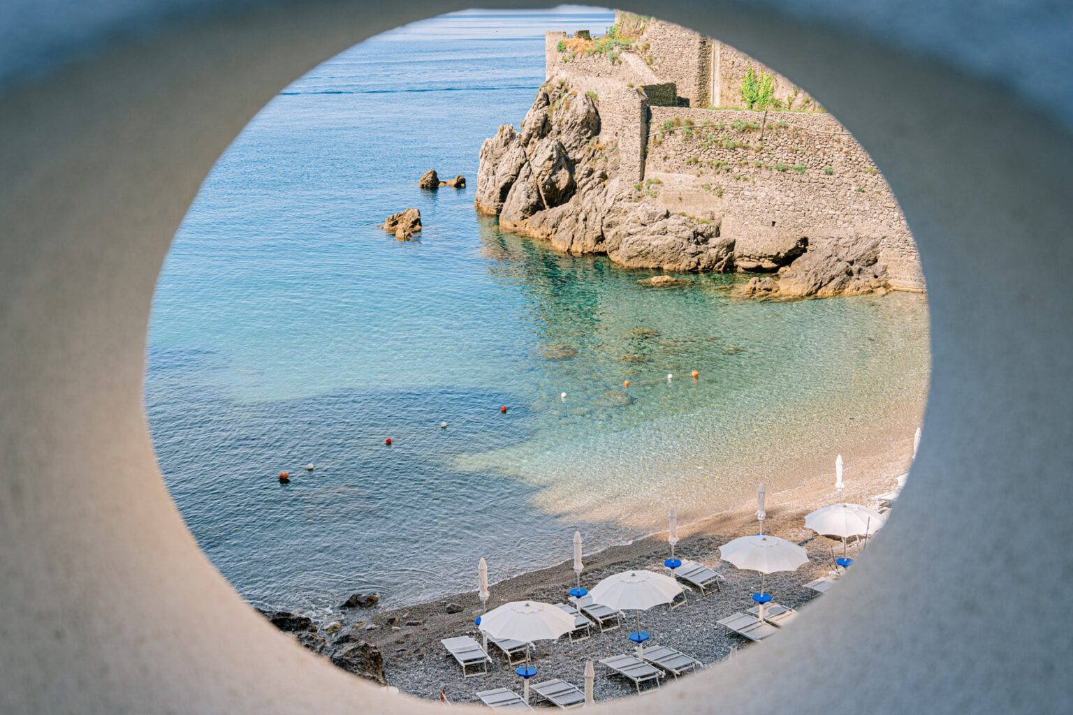 View of a serene beach framed by a circular opening, showcasing clear blue water, rocky shoreline, and sun loungers on the sand. | Only on SopranoVillas