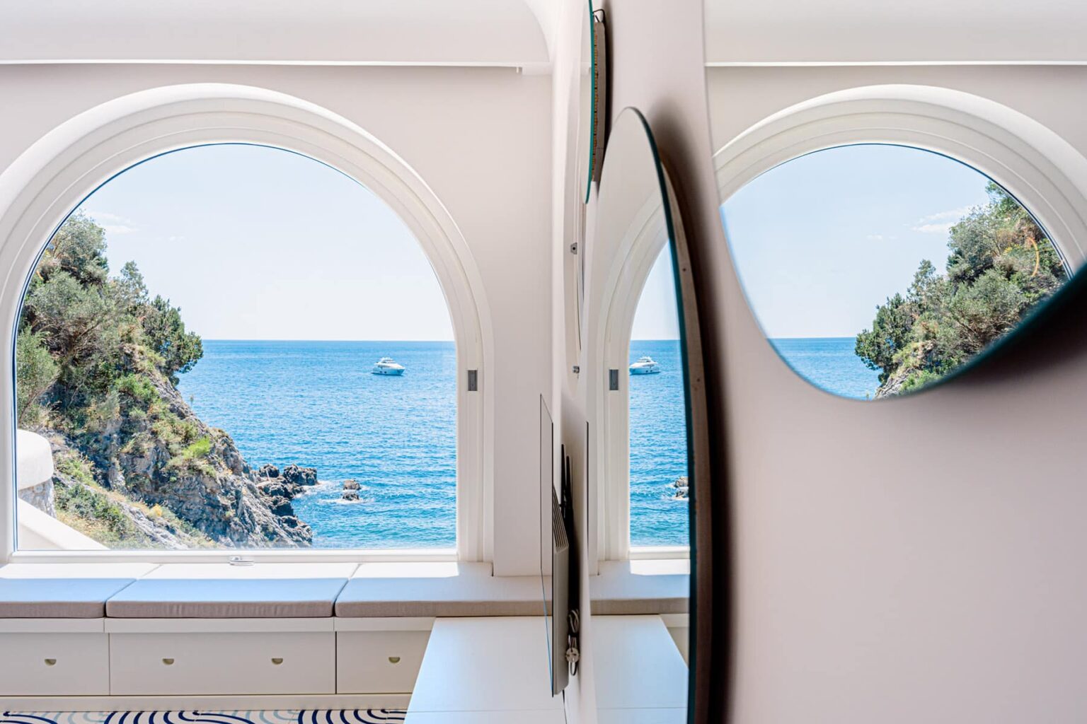 View of a serene coastal landscape through arched windows, showcasing the blue sea, distant boats, and lush greenery, enhancing the tranquil interior setting. | Only on SopranoVillas