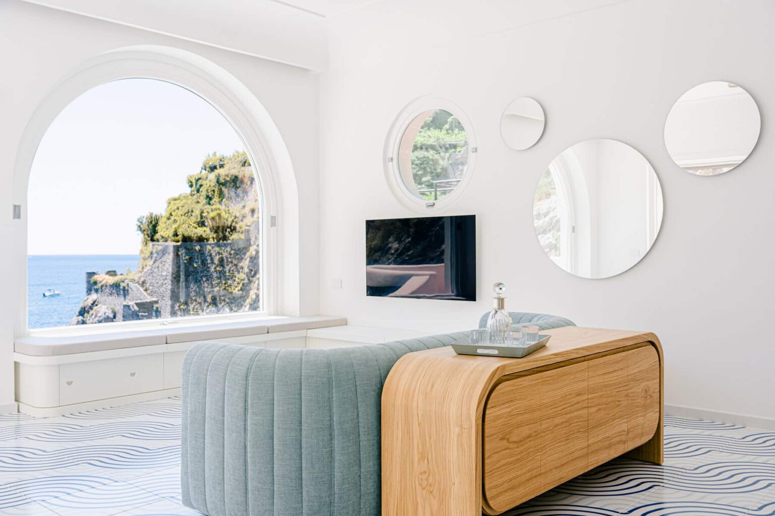 Modern living room with a curved blue sofa, wooden coffee table, and a large arched window showcasing a coastal view. Mirrors adorn the white walls, and a flat-screen TV is mounted for entertainment. The floor features a wave-patterned design, adding a touch of elegance to the space. | Only on SopranoVillas