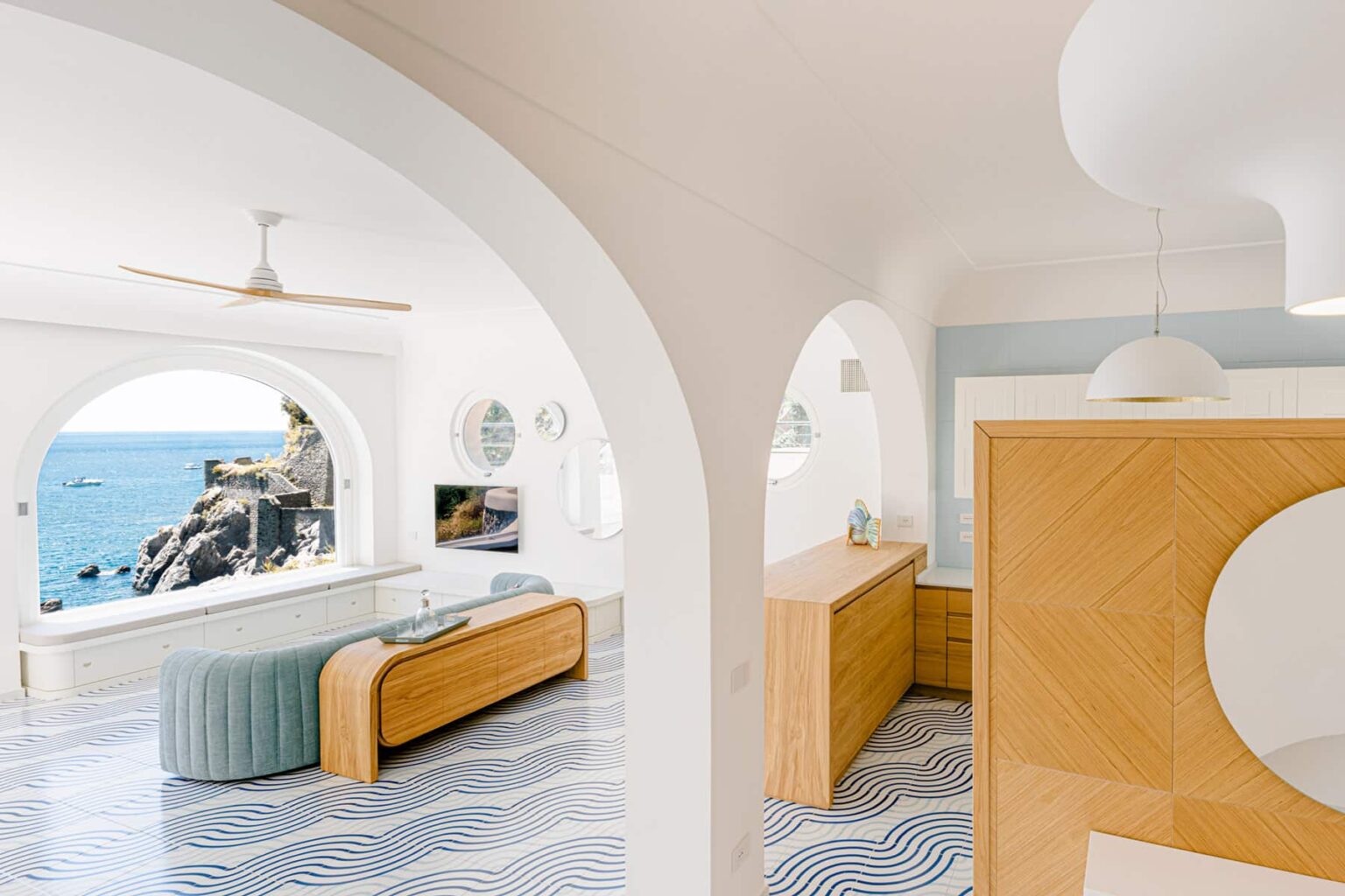 Modern coastal living room featuring arched doorways, a wooden sideboard, and a comfortable sofa with ocean views. The space is decorated in light colors with patterned tile flooring and contemporary light fixtures. | Only on SopranoVillas
