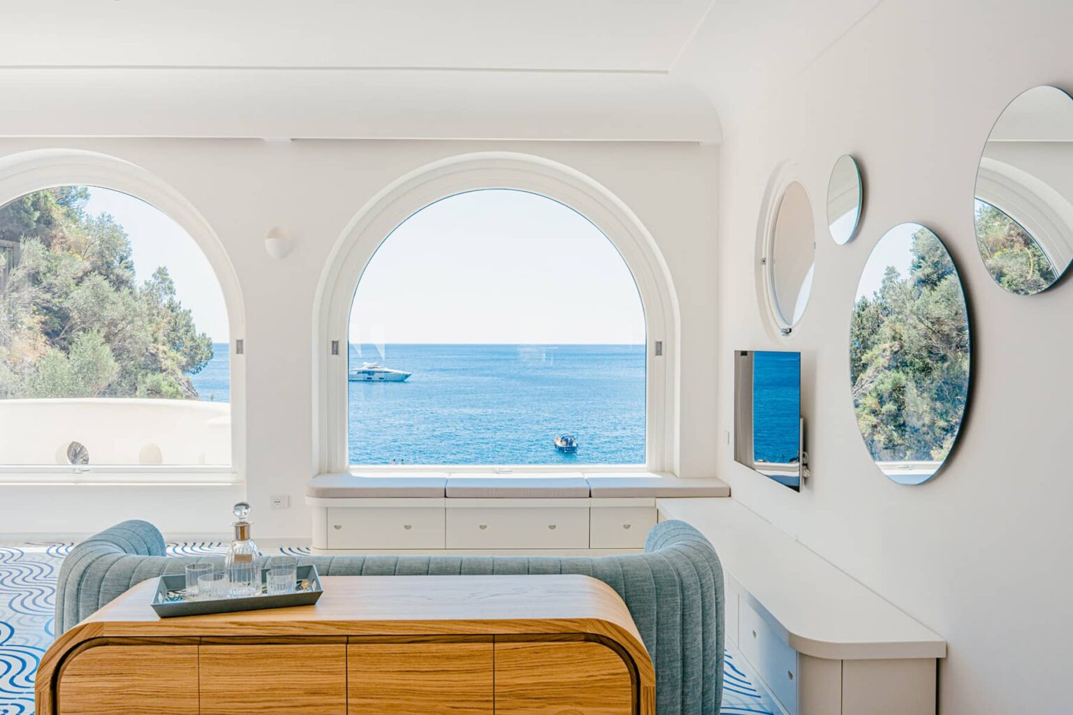 Bright and airy interior of a coastal home featuring large arch windows with a view of the ocean, modern furniture, and decorative mirrors on the walls. | Only on SopranoVillas