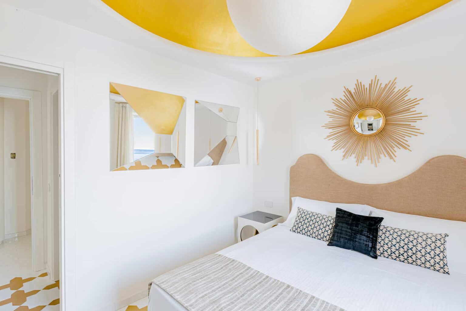Bright and modern bedroom featuring a yellow ceiling, stylish sunburst mirror, and minimalist decor. The room includes a cozy bed with decorative pillows and sleek side tables, creating a welcoming atmosphere. | Only on SopranoVillas