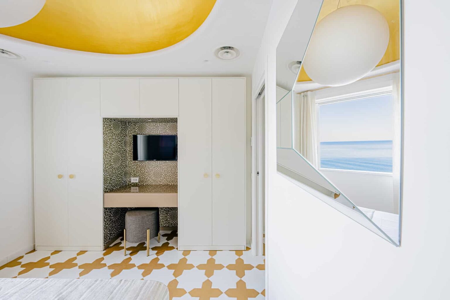 Modern interior design of a bright bedroom featuring a yellow ceiling, a minimalist wardrobe, a small desk area with a TV, and a large window offering a scenic ocean view. The floor is decorated with a stylish geometric pattern. | Only on SopranoVillas