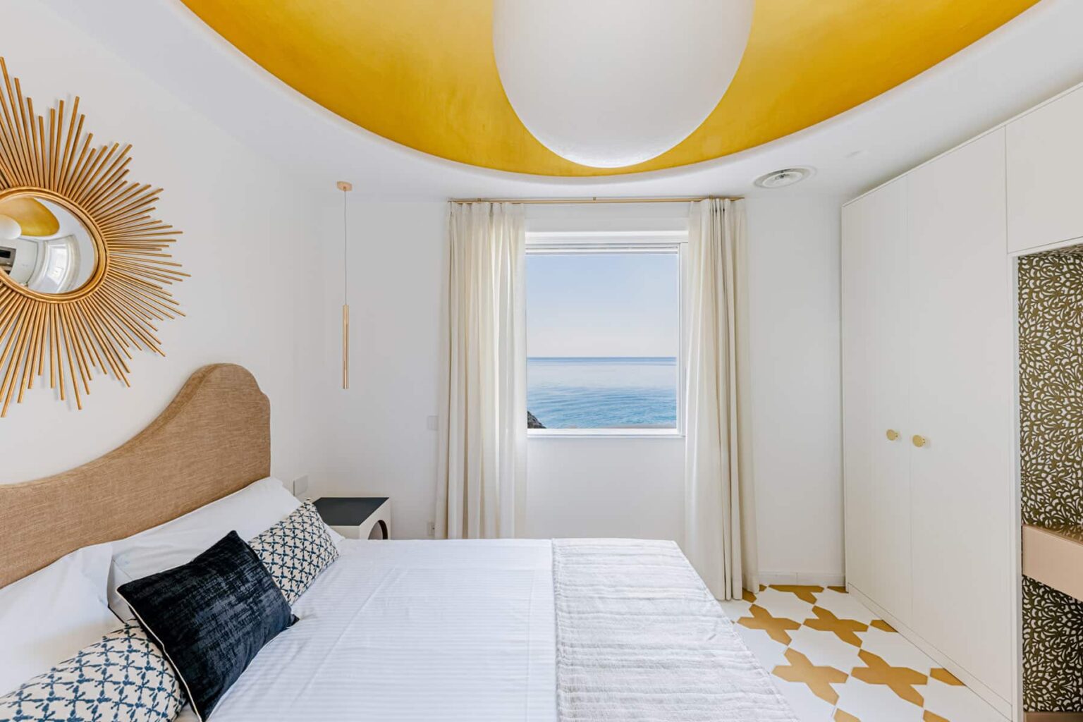 Bright and modern bedroom with a yellow ceiling, ocean view, and stylish decor. Features a plush bed with decorative pillows, large windows with sheer curtains, and contemporary furniture. Ideal for a relaxing getaway. | Only on SopranoVillas