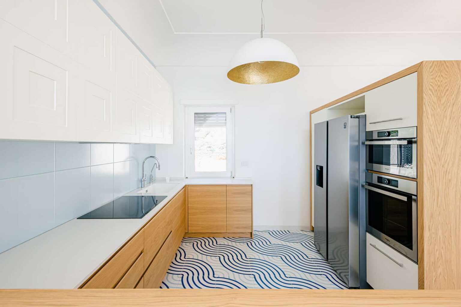 Modern kitchen with light wood cabinetry, sleek white countertops, and stainless steel appliances. The design features a unique wave-patterned floor and a stylish pendant light, creating a bright and inviting atmosphere. | Only on SopranoVillas