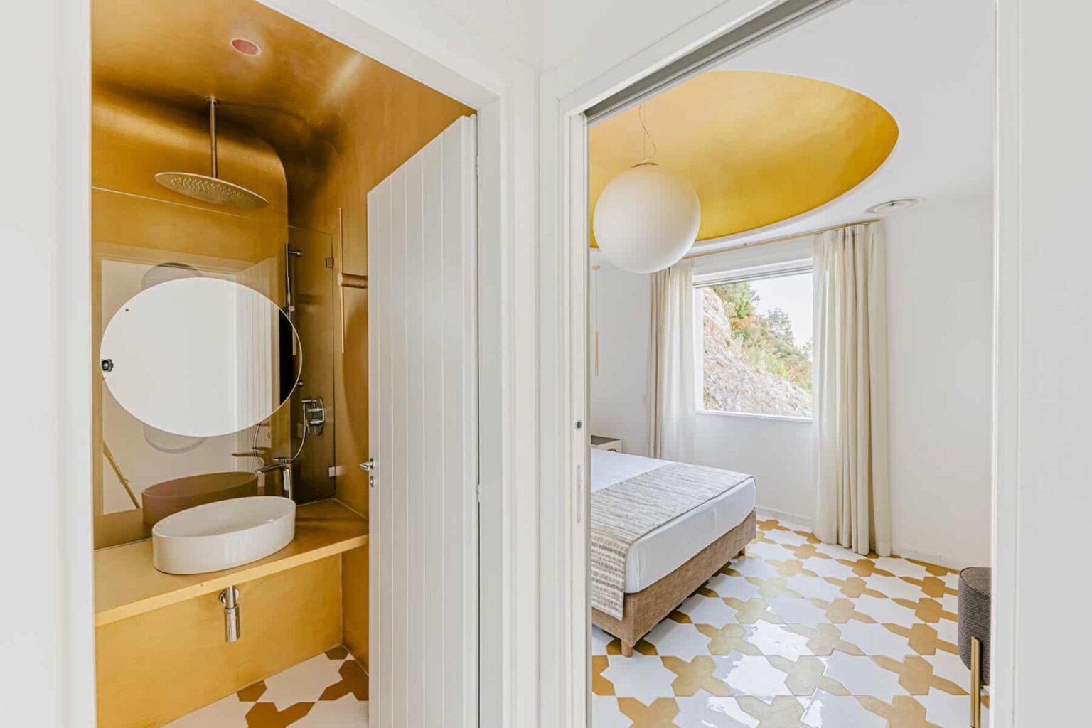 Modern interior view showcasing a stylish bathroom with a circular mirror and a sleek sink, leading into a well-lit bedroom featuring a light-colored bed and large window, all set against a unique patterned floor. | Only on SopranoVillas