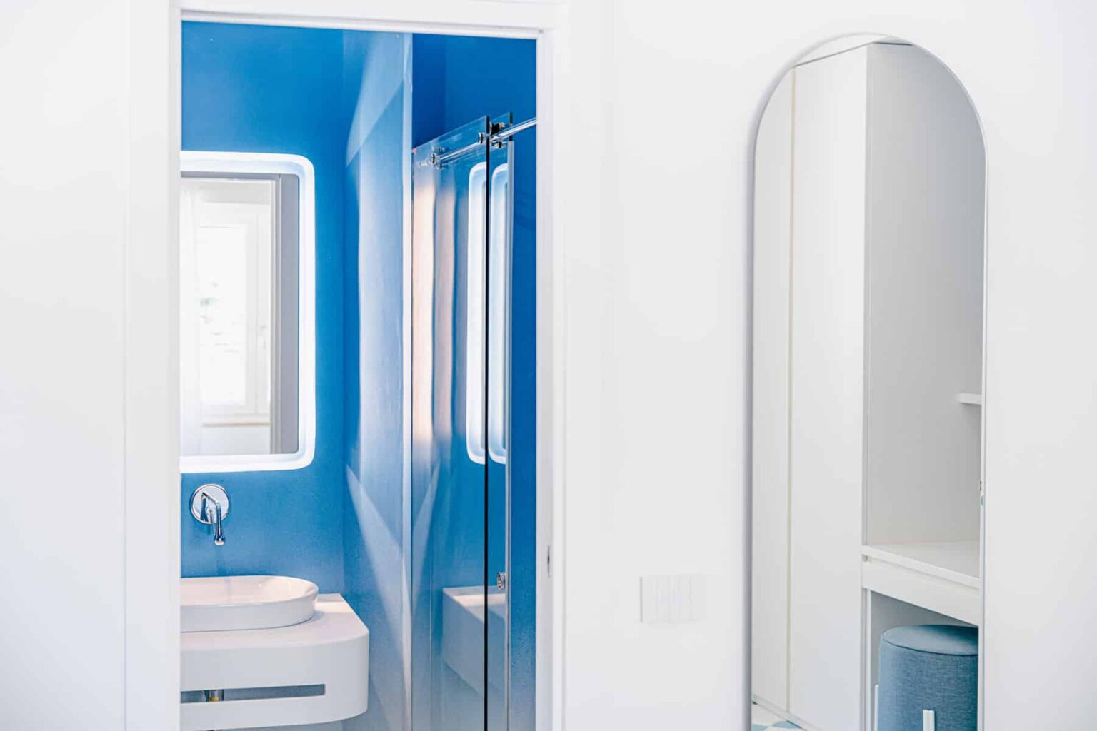 Modern bathroom interior featuring a blue accent wall, a sleek white vanity with a sink, a glass shower enclosure, and a round mirror. Brightly lit with natural light, creating a spacious and inviting atmosphere. | Only on SopranoVillas