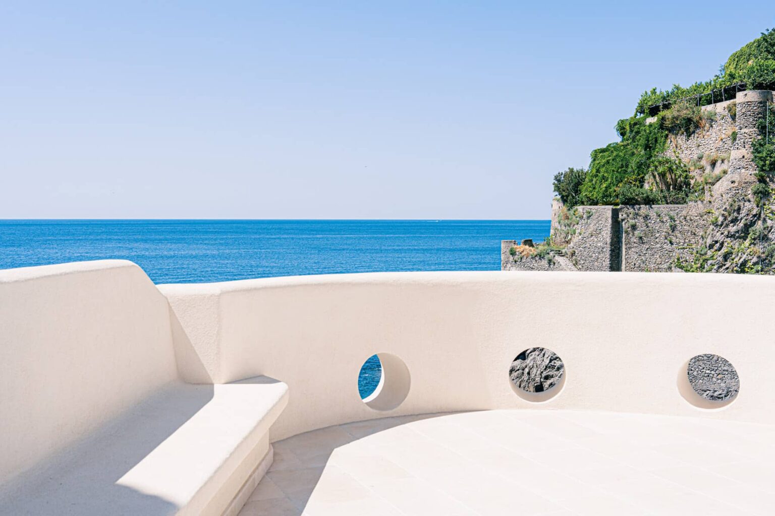 Modern terrace overlooking a serene blue ocean, featuring a white curved bench and circular cutouts in the wall, surrounded by lush greenery and rocky cliffs. Perfect for relaxation and enjoying coastal views. | Only on SopranoVillas