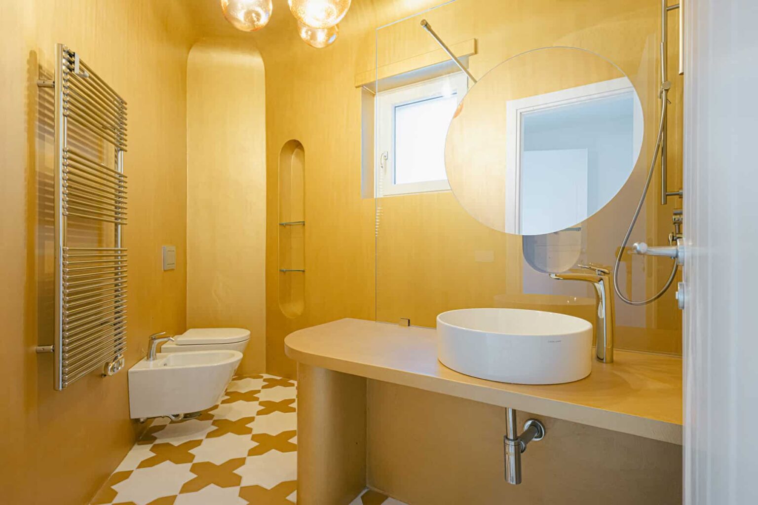 Modern bathroom design featuring a yellow color scheme, circular mirror, sleek countertop with a round sink, and stylish tile flooring. The space includes a bidet, towel radiator, and natural light from a window, creating a bright and inviting atmosphere. | Only on SopranoVillas