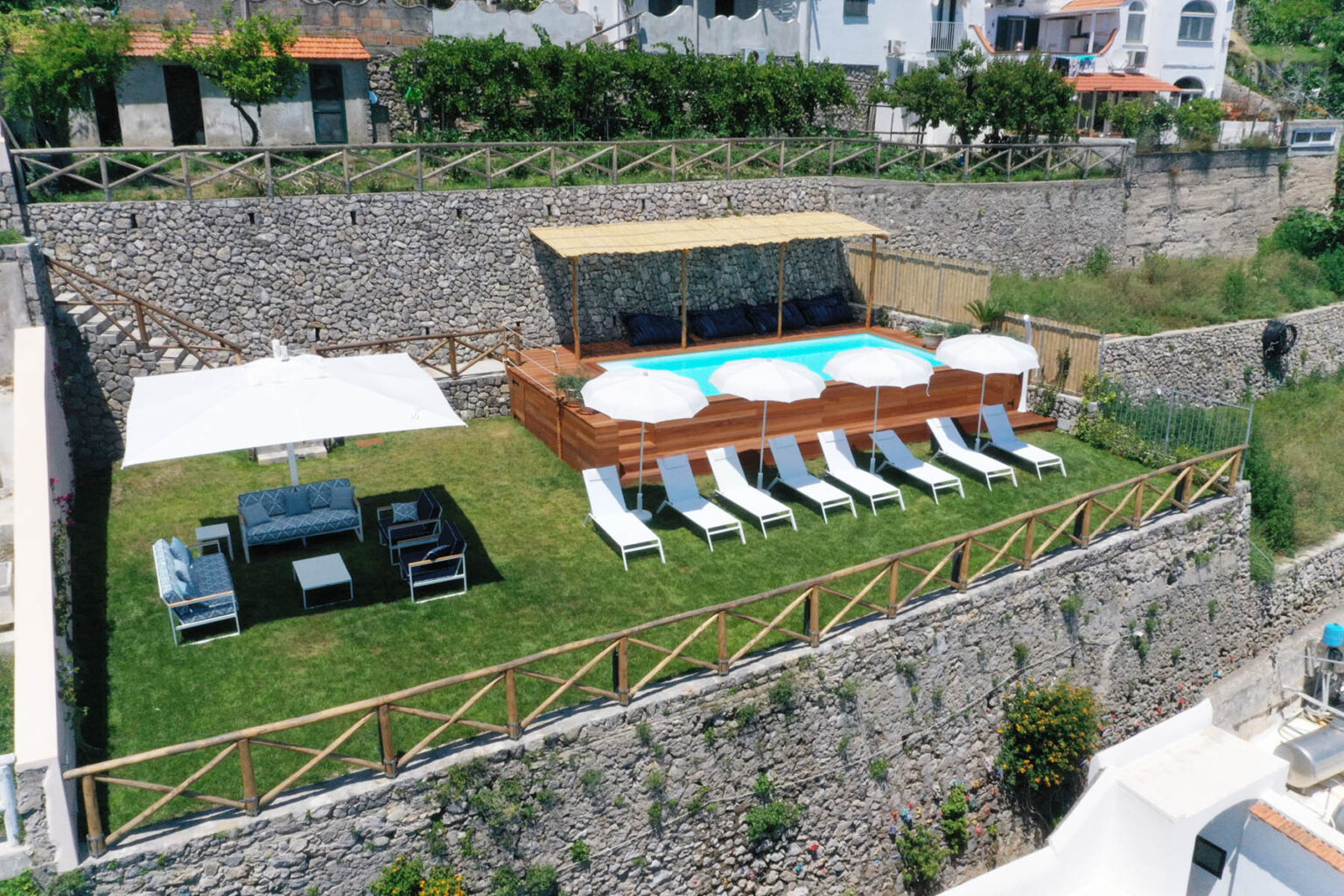 Aerial view of a luxurious outdoor pool area featuring a well-maintained lawn, sun loungers, and a shaded seating arrangement, surrounded by stone walls and greenery, ideal for relaxation and leisure. | Only on SopranoVillas