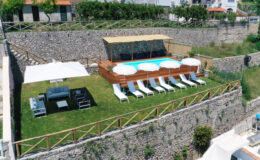 Aerial view of a luxurious outdoor pool area featuring a well-maintained lawn, sun loungers, and a shaded seating arrangement, surrounded by stone walls and greenery, ideal for relaxation and leisure.