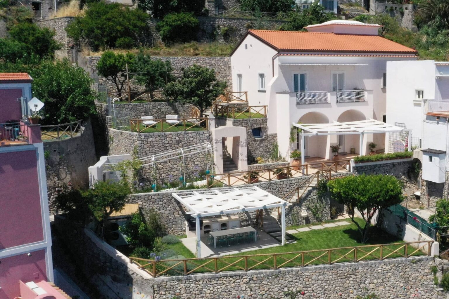 Aerial view of a Mediterranean villa featuring a spacious garden, outdoor seating areas, and stone walls, surrounded by lush greenery and a clear blue sky. | Only on SopranoVillas
