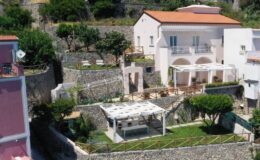 Aerial view of a Mediterranean villa featuring a spacious garden, outdoor seating areas, and stone walls, surrounded by lush greenery and a clear blue sky.