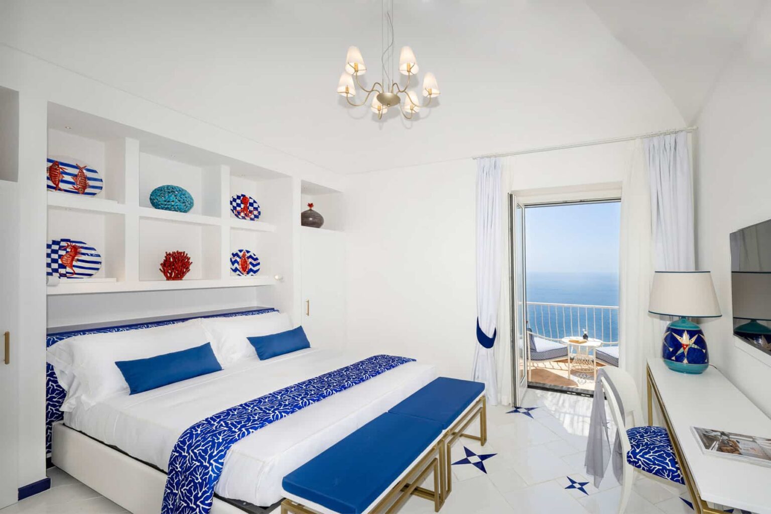 Bright and airy hotel room featuring a king-sized bed with blue and white bedding, modern decor, and a stunning ocean view through large windows. The room includes a stylish chandelier, decorative shelves, and a workspace with a desk. Ideal for a relaxing getaway. | Only on SopranoVillas