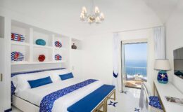 Bright and airy hotel room featuring a king-sized bed with blue and white bedding, modern decor, and a stunning ocean view through large windows. The room includes a stylish chandelier, decorative shelves, and a workspace with a desk. Ideal for a relaxing getaway.