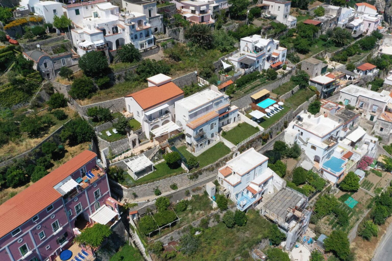Aerial view of a hillside residential area featuring white houses with orange roofs, green gardens, and a swimming pool, surrounded by lush vegetation. | Only on SopranoVillas